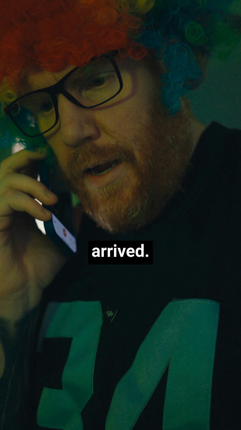 A man with a red beard and glasses wears a colorful curly clown wig while talking on a smartphone. The word "arrived." is displayed in bold white text over a black background near the bottom center of the image.