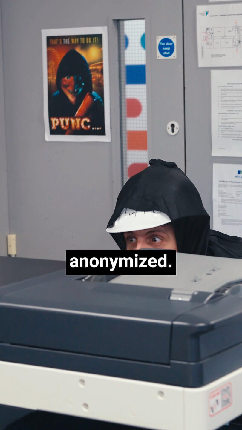 A person wearing a black hood and white construction helmet crouches behind an office printer, peeking over it. The word “anonymised.” appears in bold white text on a black background near the bottom centre.