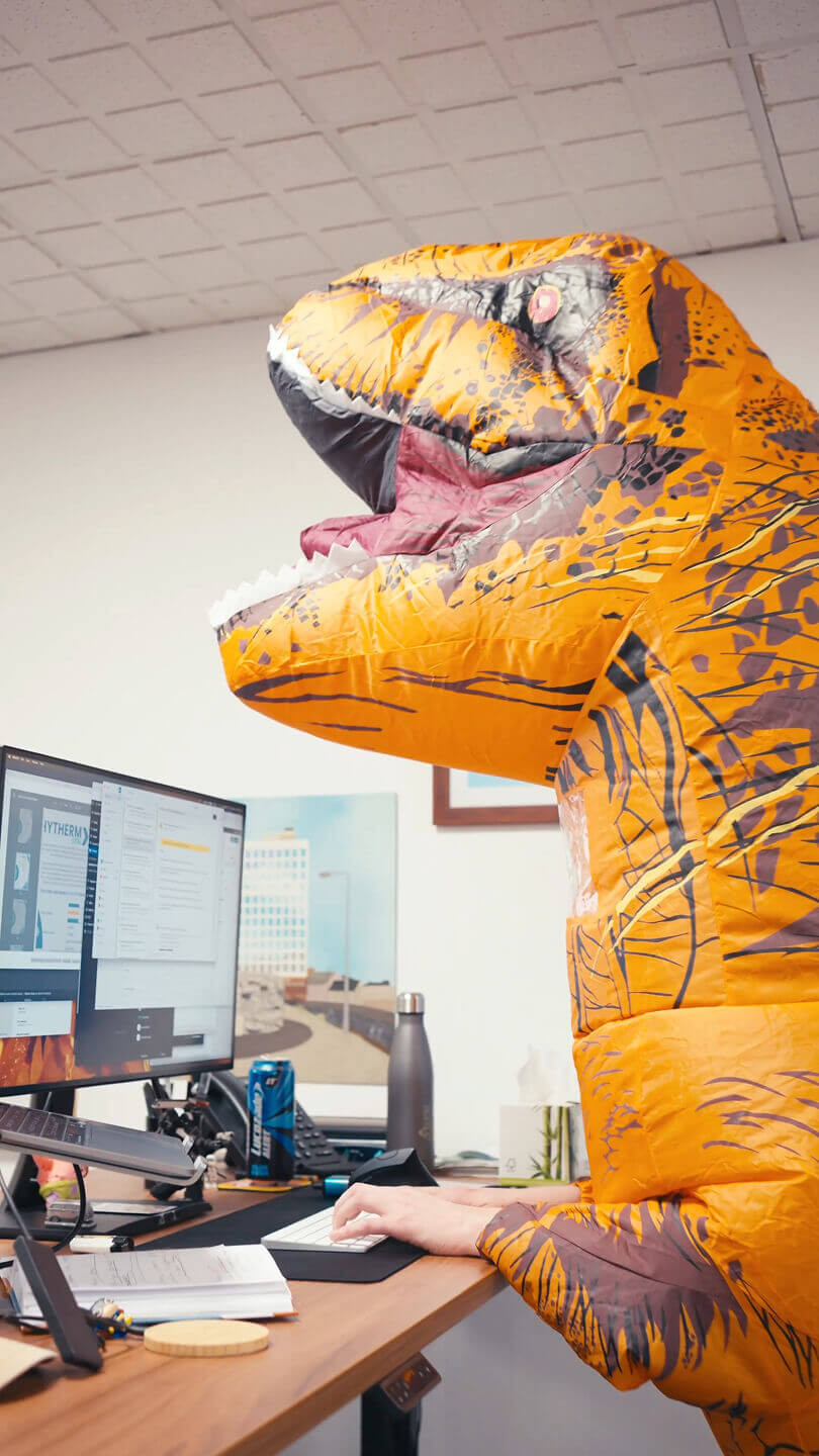A man in a dinosaur outfit in an office stood at a desk typing on a keyboard. The desk is cluttered with refreshments and personal items.