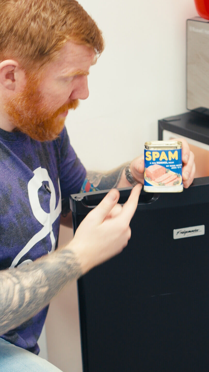 A man knelt down by an open fridge. One arm is resting on the fridge whilst the other is pointing to it retrieving and pointing to a can of ‘Spam”.