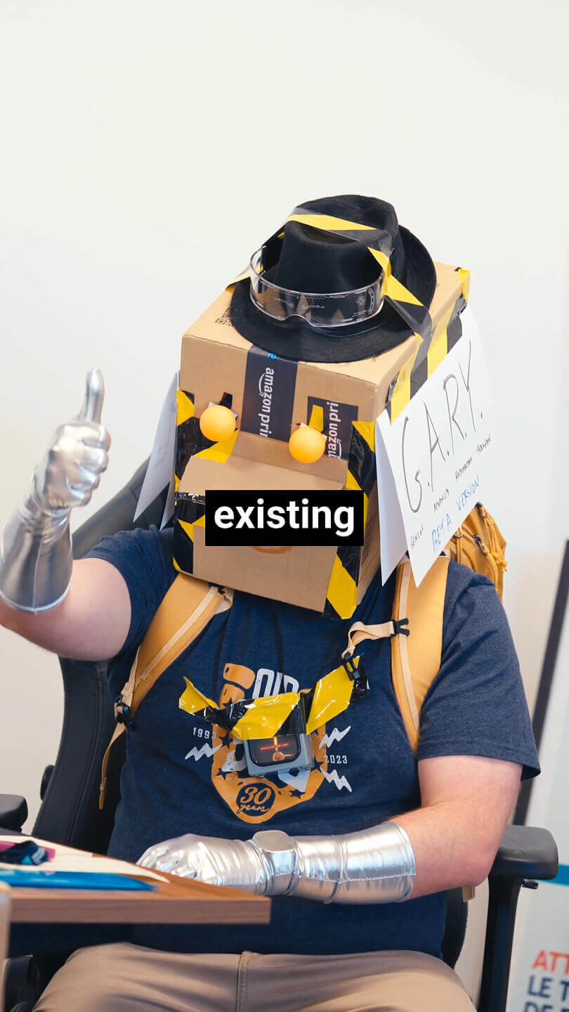 A man dressed in a crudely made robot costume with a cardboard box for a head and tight silver arm length gloves on. His right arm is raised displaying a thumbs up.