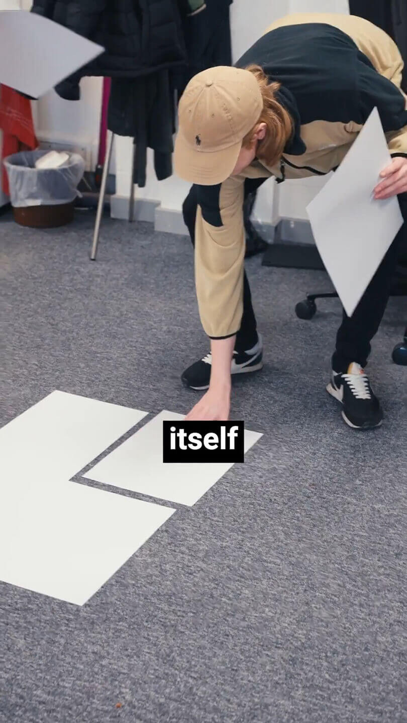 A man in an office environment crouching down and placing large sheets of paper side by side on the floor.