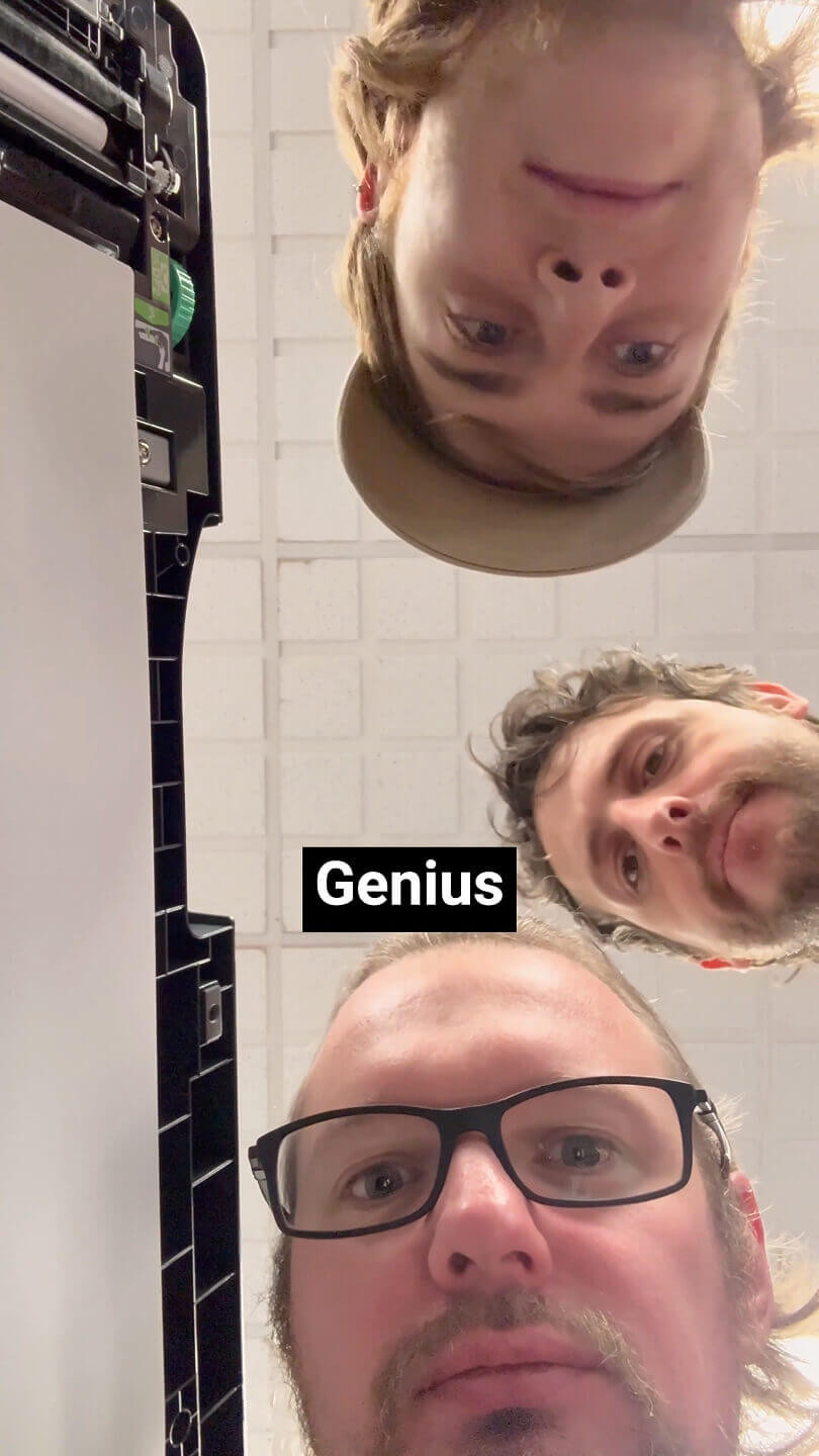 3 Men stood over an officer printer looking down into the camera as if they are looking into the printer.