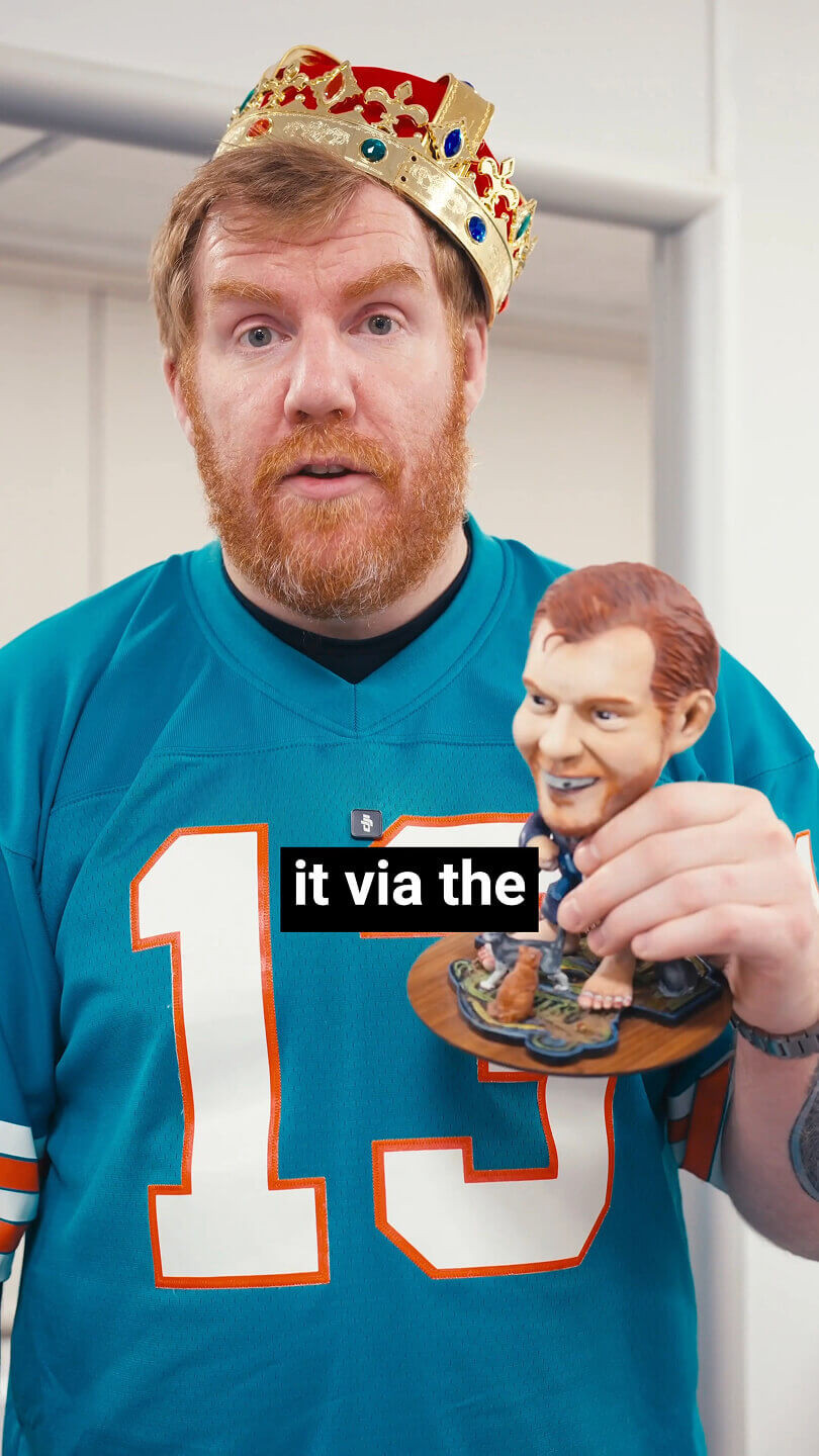 A man with a plastic crown sitting slanted upon his head. In his hand he is holding a small plastic version of himself.