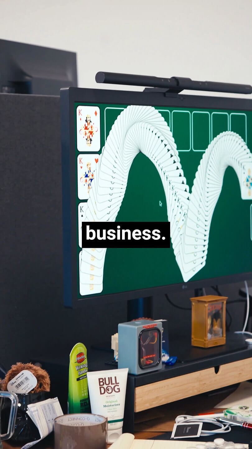 Computer screen shows Solitaire win animation with the word “business.” overlaid; cluttered desk in foreground.